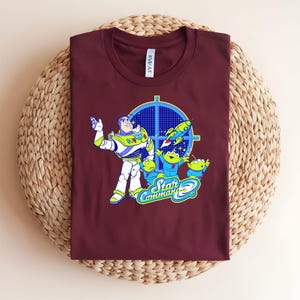 May include: A maroon t-shirt with a graphic of Buzz Lightyear and the Little Green Men from Toy Story. The design includes the words "Star Command" in a blue and green font, with a target and rocket ship.