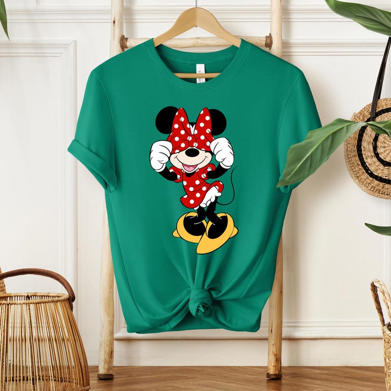 Pu&ograve; includere: Una maglietta verde con una grafica di Minnie Mouse. La grafica mostra Minnie Mouse con un vestito a pois rossi, fiocco abbinato e scarpe gialle. La maglietta &egrave; appesa a un supporto di legno.