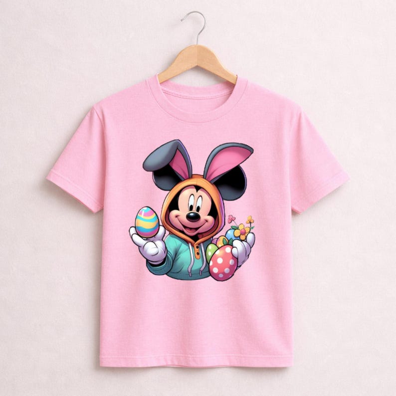 May include: Pink t-shirt featuring a cartoon Mickey Mouse character dressed as the Easter Bunny. Mickey is holding colorful Easter eggs and flowers. The shirt has a crew neck and short sleeves.
