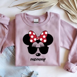 Może przedstawiać: Jasnor&oacute;żowa bluza z nadrukiem Myszki Minnie w okularach przeciwsłonecznych i czerwoną kokardą w białe grochy. Słowo "mommy" jest wydrukowane poniżej na czarno. Bluza jest pokazana z niebieskimi dżinsami.