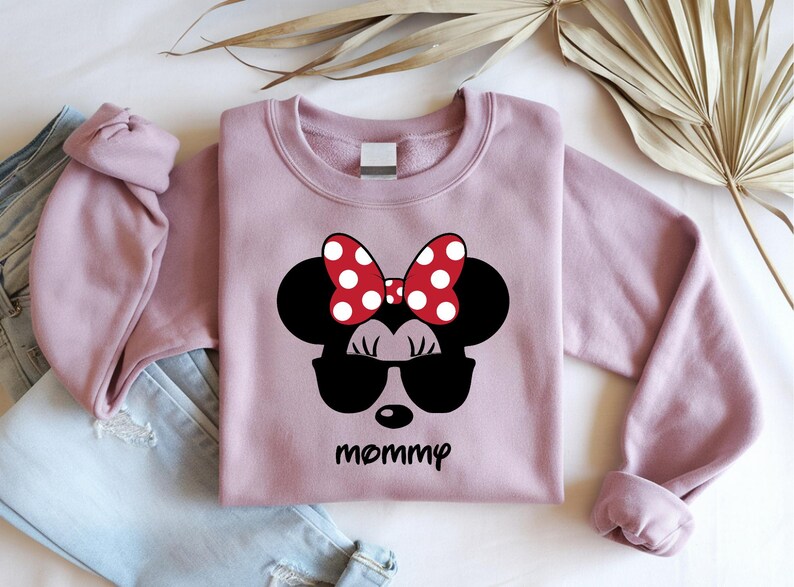 Może przedstawiać: Jasnor&oacute;żowa bluza z nadrukiem Myszki Minnie w okularach przeciwsłonecznych i czerwoną kokardą w białe grochy. Słowo "mommy" jest wydrukowane poniżej na czarno. Bluza jest pokazana z niebieskimi dżinsami.