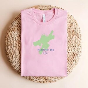 May include: A pale pink t-shirt featuring a green graphic and the text "Reach for the sky!" in blue and green. The shirt is folded on a woven, circular surface.
