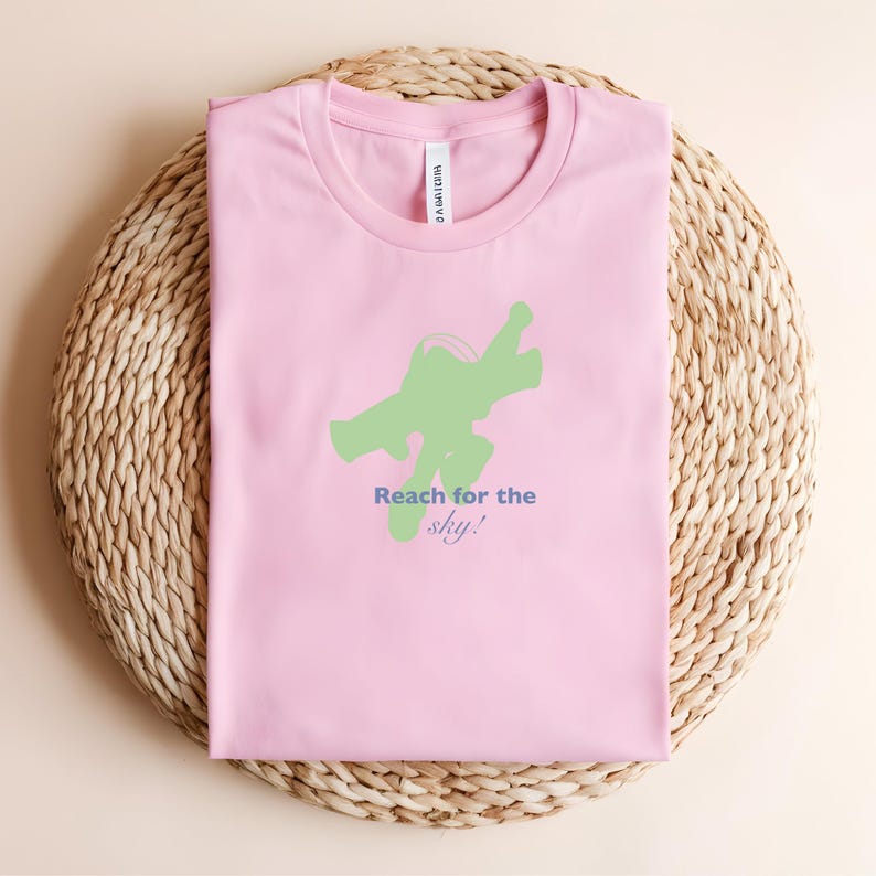 May include: A pale pink t-shirt featuring a green graphic and the text "Reach for the sky!" in blue and green. The shirt is folded on a woven, circular surface.