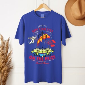 May include: Royal blue t-shirt featuring a graphic design with the text "KEEP YOUR EYES ON THE PRIZE" and the Pizza Planet logo. The design includes cartoon aliens, Buzz Lightyear, and a pizza slice.