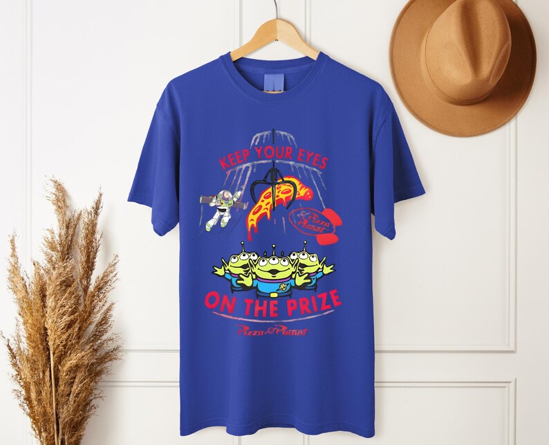 May include: Royal blue t-shirt featuring a graphic design with the text "KEEP YOUR EYES ON THE PRIZE" and the Pizza Planet logo. The design includes cartoon aliens, Buzz Lightyear, and a pizza slice.