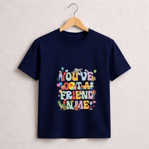 May include: Navy blue t-shirt with the phrase "YOU'VE GOT A FRIEND IN ME" in colorful, playful lettering. The design includes cartoon characters and balloons. The shirt is hanging on a wooden hanger.