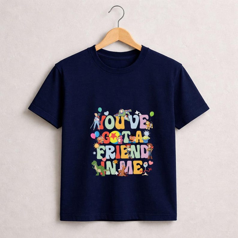 May include: Navy blue t-shirt with the phrase "YOU'VE GOT A FRIEND IN ME" in colorful, playful lettering. The design includes cartoon characters and balloons. The shirt is hanging on a wooden hanger.
