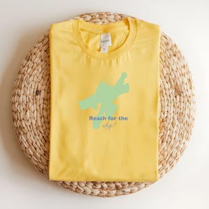 May include: A yellow t-shirt featuring a light green abstract shape and the blue text "Reach for the sky!". The shirt is folded and placed on a circular woven mat.