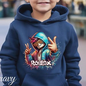 Puede incluir: Sudadera con capucha azul marino con un gr&aacute;fico colorido de un personaje con gafas de sol haciendo el signo de la paz. La palabra "ROBLOX" se muestra en una fuente vibrante y multicolor. La sudadera tiene una capucha con cord&oacute;n y un bolsillo delantero.