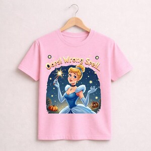 May include: Pink t-shirt featuring a cartoon illustration of Cinderella holding a wand. The text "Oops! Wrong Spell..." is above the image. Cinderella is wearing a blue dress and gloves, with a pumpkin and carriage in the background.