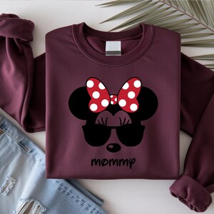 Może przedstawiać: Bordowa bluza z czarnym wzorem Myszki Minnie w okularach przeciwsłonecznych i czerwoną kokardą w grochy. Słowo "mommy" jest napisane czarną czcionką poniżej. Bluza jest złożona z podwiniętymi rękawami.