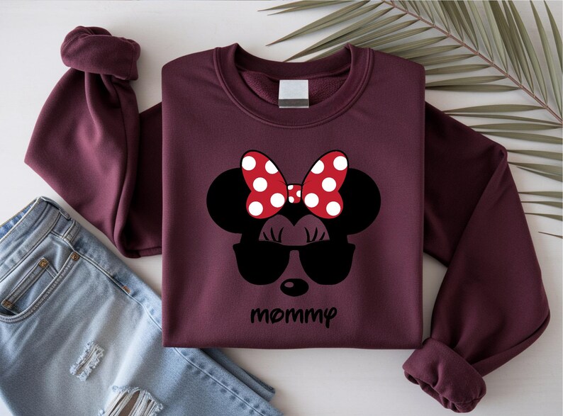 Może przedstawiać: Bordowa bluza z czarnym wzorem Myszki Minnie w okularach przeciwsłonecznych i czerwoną kokardą w grochy. Słowo "mommy" jest napisane czarną czcionką poniżej. Bluza jest złożona z podwiniętymi rękawami.