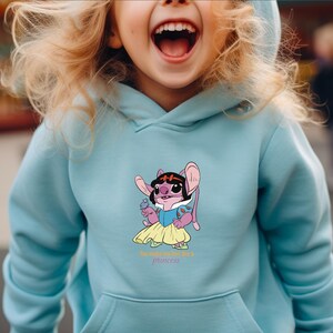 May include: Light blue hooded sweatshirt featuring a cartoon character dressed as a princess. The character is pink with large ears, wearing a black headband, blue dress, and yellow skirt. The text "the cutest little princess" is below the graphic.