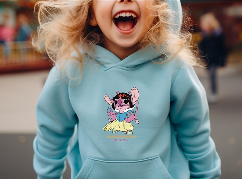 May include: Light blue hooded sweatshirt featuring a cartoon character dressed as a princess. The character is pink with large ears, wearing a black headband, blue dress, and yellow skirt. The text "the cutest little princess" is below the graphic.