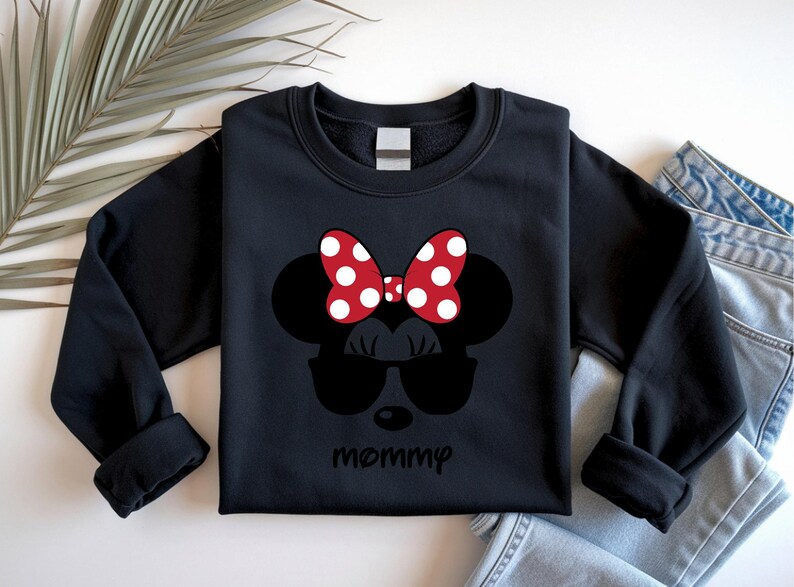 Może przedstawiać: Czarna bluza z nadrukiem Myszki Minnie w okularach przeciwsłonecznych i czerwoną kokardą w białe grochy. Poniżej nadrukowano słowo "mommy". Bluza jest prezentowana na białej powierzchni.