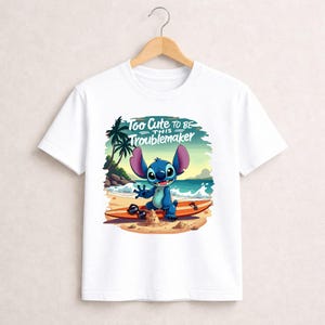 Pu&ograve; includere: T-shirt bianca con un personaggio dei cartoni animati su una tavola da surf sulla spiaggia. Il personaggio blu ha grandi orecchie e sorride. Il testo "Too Cute to Be This Troublemaker" &egrave; sopra il personaggio.