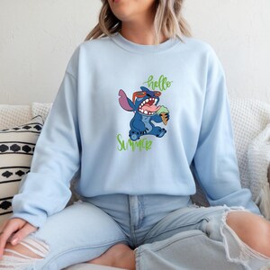 Puede incluir: Sudadera azul claro con un personaje de dibujos animados, Stitch, con gafas de sol y un helado. Las palabras "hello" y "Summer" est&aacute;n escritas en verde. La sudadera es de color azul claro liso.