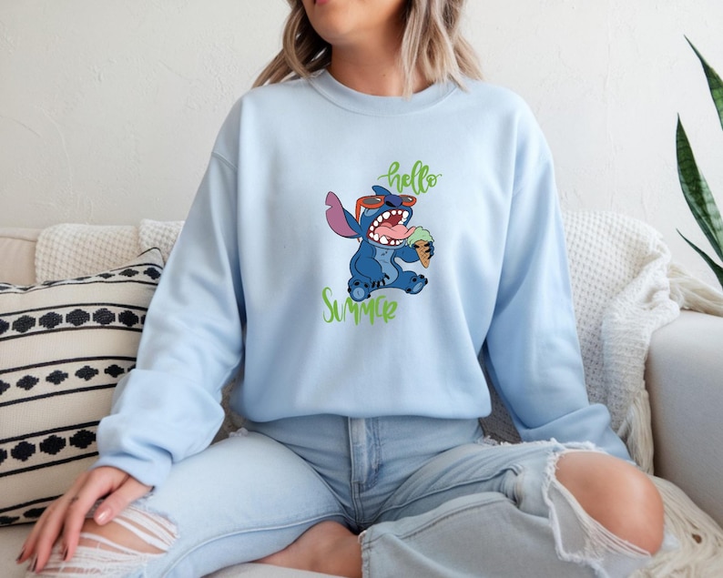 Puede incluir: Sudadera azul claro con un personaje de dibujos animados, Stitch, con gafas de sol y un helado. Las palabras "hello" y "Summer" est&aacute;n escritas en verde. La sudadera es de color azul claro liso.