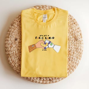 May include: Yellow t-shirt with the text "you got a FRIEND in me" and a graphic of two hands forming a heart. One hand is human, the other is a robot hand. The shirt is on a woven, circular surface.