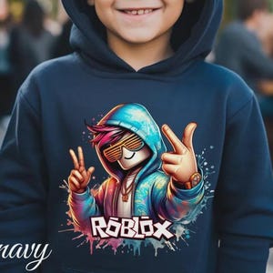 Puede incluir: Sudadera con capucha azul marino con un gr&aacute;fico colorido de un personaje de Roblox con gafas de sol y haciendo el signo de la paz. La palabra "ROBLOX" est&aacute; impresa en blanco y rosa. La palabra "navy" est&aacute; escrita en cursiva en la parte inferior izquierda.