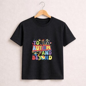 Op de afbeelding: Zwart T-shirt met een kleurrijke afbeelding met de tekst "TO AUTISM AND BEYOND". Het ontwerp bevat stripfiguren en een raket. Het T-shirt hangt aan een houten hanger.