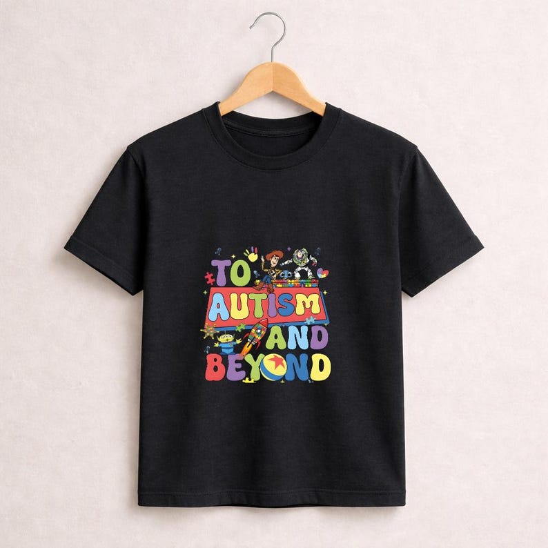 Op de afbeelding: Zwart T-shirt met een kleurrijke afbeelding met de tekst "TO AUTISM AND BEYOND". Het ontwerp bevat stripfiguren en een raket. Het T-shirt hangt aan een houten hanger.