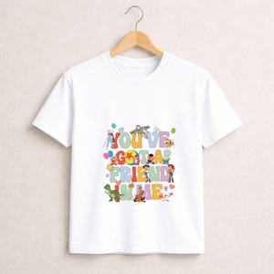 May include: White t-shirt featuring the phrase "YOU'VE GOT A FRIEND IN ME" in colorful, playful lettering. The design includes cartoon characters from a popular animated movie, with a wooden hanger in the background.