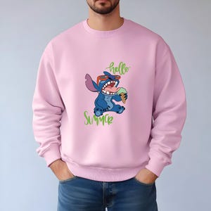 Puede incluir: Sudadera rosa claro con un personaje de dibujos animados azul con gafas de sol y comiendo helado. Las palabras "hello" y "Summer" est&aacute;n escritas en verde. La sudadera es de color rosa liso.