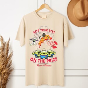 May include: Tan t-shirt featuring a graphic from the movie Toy Story. The design includes the text "KEEP YOUR EYES ON THE PRIZE" with images of the Little Green Men, Buzz Lightyear, and a pizza slice. The Pizza Planet logo is also visible.