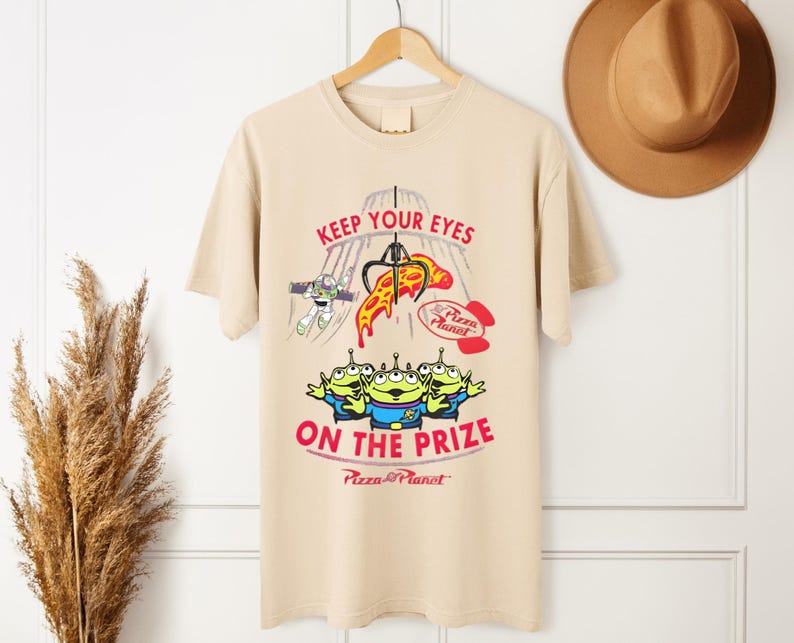 May include: Tan t-shirt featuring a graphic from the movie Toy Story. The design includes the text "KEEP YOUR EYES ON THE PRIZE" with images of the Little Green Men, Buzz Lightyear, and a pizza slice. The Pizza Planet logo is also visible.