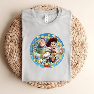 Puede incluir: Una camiseta gris claro con un gr&aacute;fico circular de Buzz Lightyear y Woody de Toy Story. El gr&aacute;fico est&aacute; rodeado por un borde azul con estrellas amarillas y las palabras "TOY STORY".