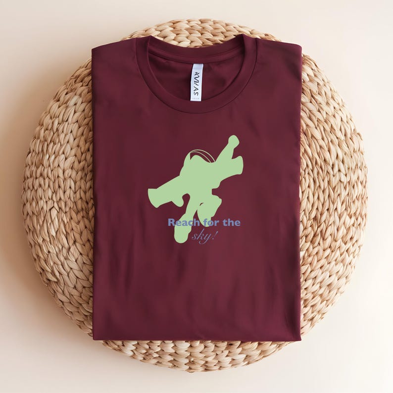 May include: A maroon t-shirt featuring a light green graphic of a character in flight. The text "Reach for the sky!" is printed below the graphic in blue and green. The shirt is folded and placed on a woven, circular surface.