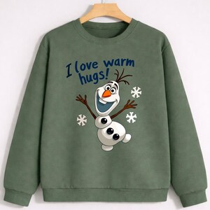 May include: Green sweatshirt featuring a cartoon Olaf character with the text "I love warm hugs!" in blue. The design includes snowflakes. The long-sleeved top is made of a soft material.
