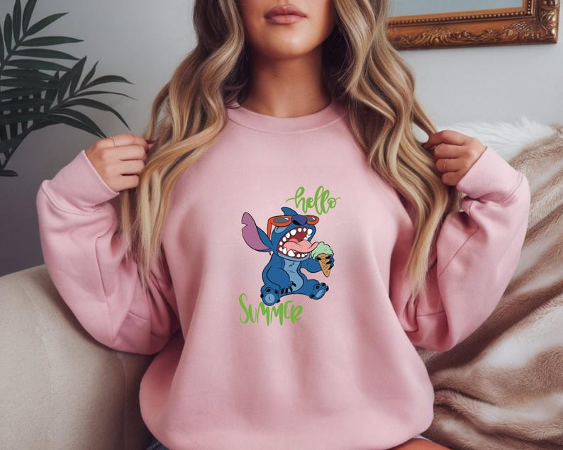 Puede incluir: Una sudadera rosa claro con un personaje de dibujos animados azul que lleva gafas de sol y sostiene un cono de helado. Las palabras "hello summer" est&aacute;n escritas en verde arriba y abajo del personaje.