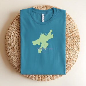 May include: Teal crew neck t-shirt with a light green space ranger graphic and the text "Reach for the sky!" in purple. The shirt is folded on a woven, circular placemat.