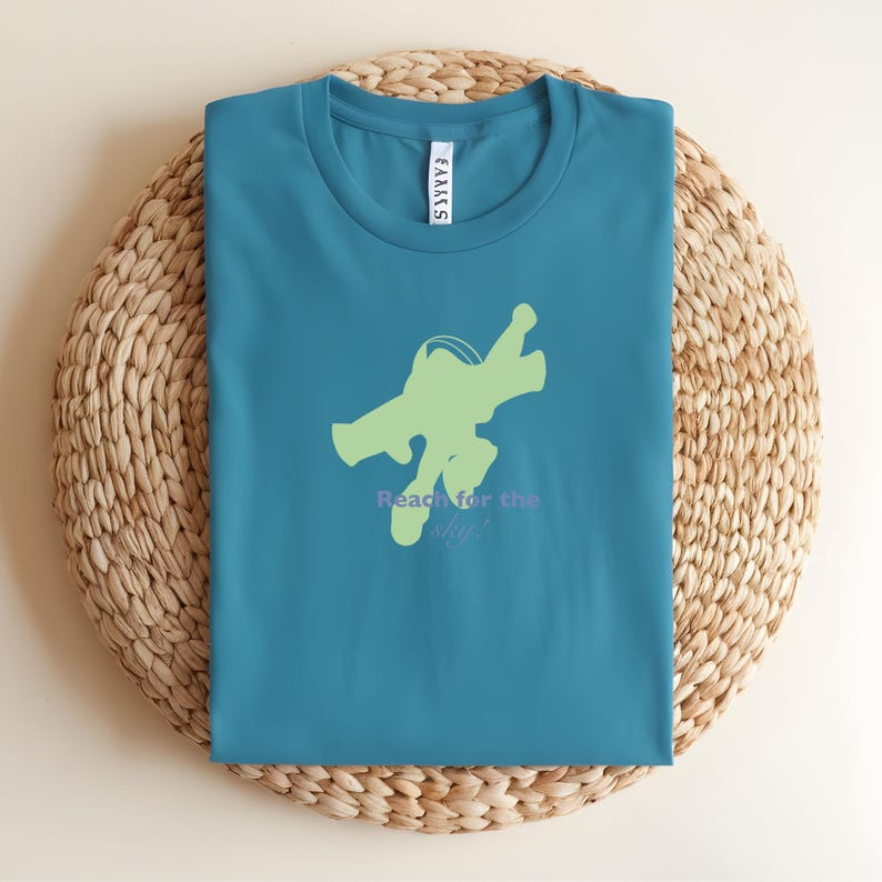 May include: Teal crew neck t-shirt with a light green space ranger graphic and the text "Reach for the sky!" in purple. The shirt is folded on a woven, circular placemat.