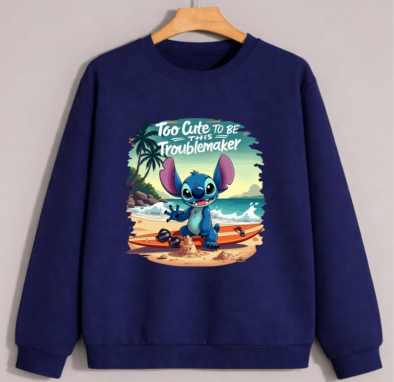 Puede incluir: Sudadera azul marino con un gr&aacute;fico de Stitch en una tabla de surf en la playa. El texto "Too Cute to be this Troublemaker" est&aacute; encima de Stitch. El dise&ntilde;o incluye palmeras, arena y olas.