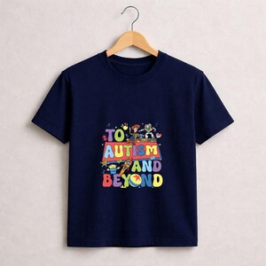 Op de afbeelding: Marineblauw T-shirt met een kleurrijke afbeelding met stripfiguren en de tekst "TO AUTISM AND BEYOND". Het ontwerp bevat een raket, aliens en andere cartoonfiguren. Het T-shirt hangt aan een houten hanger.