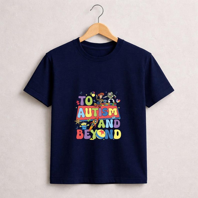 Op de afbeelding: Marineblauw T-shirt met een kleurrijke afbeelding met stripfiguren en de tekst "TO AUTISM AND BEYOND". Het ontwerp bevat een raket, aliens en andere cartoonfiguren. Het T-shirt hangt aan een houten hanger.