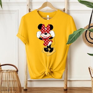 Pu&ograve; includere: T-shirt giallo senape con grafica di Minnie Mouse. Il personaggio dei cartoni animati indossa un vestito rosso a pois bianchi e un fiocco abbinato. La maglietta &egrave; annodata in vita e appesa a una gruccia di legno.