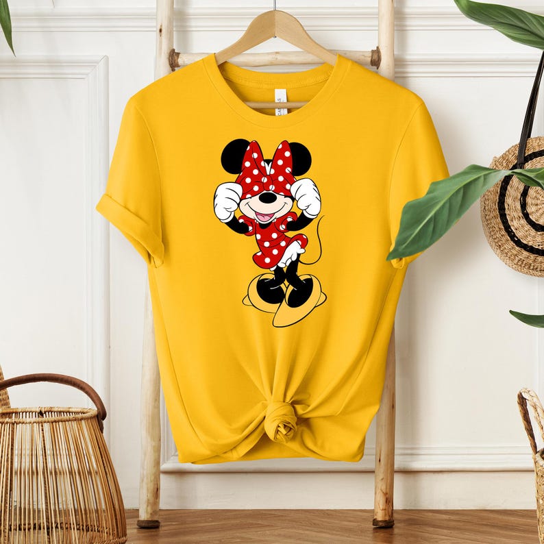 Pu&ograve; includere: T-shirt giallo senape con grafica di Minnie Mouse. Il personaggio dei cartoni animati indossa un vestito rosso a pois bianchi e un fiocco abbinato. La maglietta &egrave; annodata in vita e appesa a una gruccia di legno.