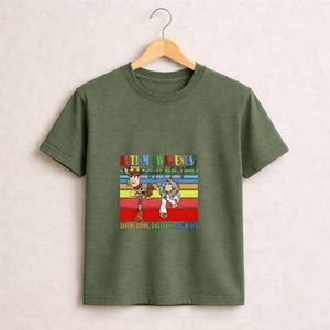 May include: Olive green t-shirt featuring a colorful graphic of Woody and Buzz Lightyear from Toy Story. The graphic includes the text "Autism Awareness And Beyond" and "Acceptance, Inclusion, Unity".
