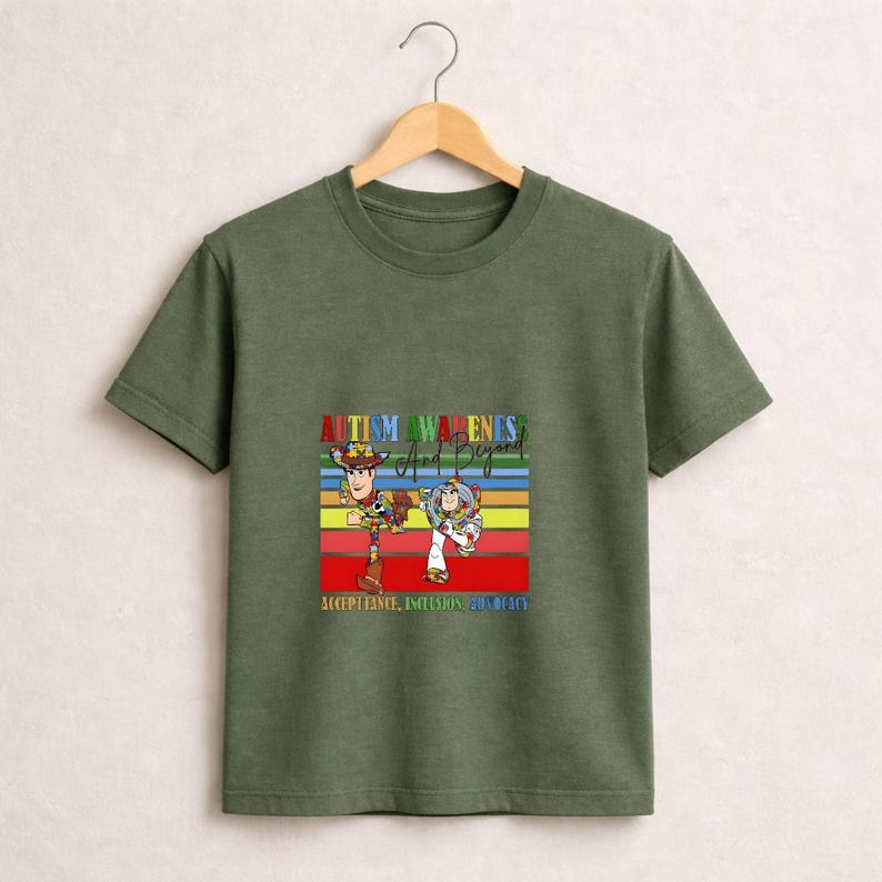 May include: Olive green t-shirt featuring a colorful graphic of Woody and Buzz Lightyear from Toy Story. The graphic includes the text "Autism Awareness And Beyond" and "Acceptance, Inclusion, Unity".