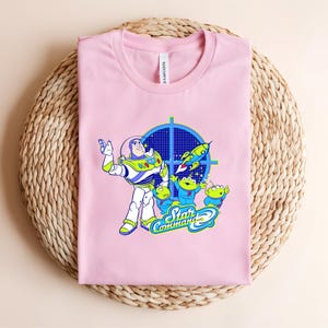 May include: A pale pink t-shirt with a graphic of Buzz Lightyear and the Little Green Men from Toy Story. The design includes a blue target, a rocket, and the words "Star Command". The shirt is folded on a woven surface.