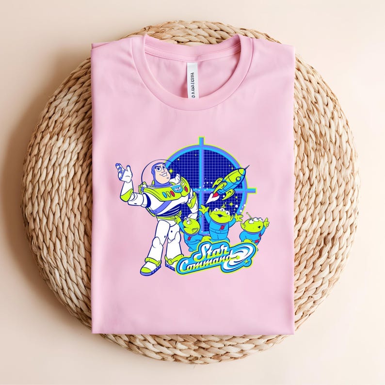 May include: A pale pink t-shirt with a graphic of Buzz Lightyear and the Little Green Men from Toy Story. The design includes a blue target, a rocket, and the words "Star Command". The shirt is folded on a woven surface.