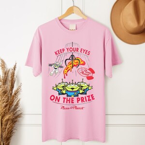 May include: Pink t-shirt featuring a graphic from the movie Toy Story. The design includes the text "KEEP YOUR EYES ON THE PRIZE" with images of the Pizza Planet aliens, Buzz Lightyear, and a pizza slice.