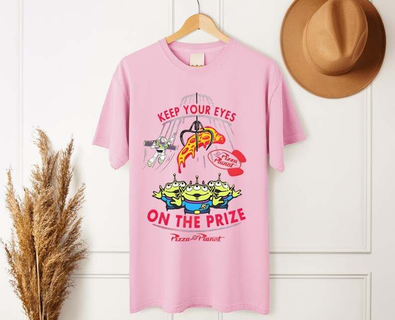 May include: Pink t-shirt featuring a graphic from the movie Toy Story. The design includes the text "KEEP YOUR EYES ON THE PRIZE" with images of the Pizza Planet aliens, Buzz Lightyear, and a pizza slice.
