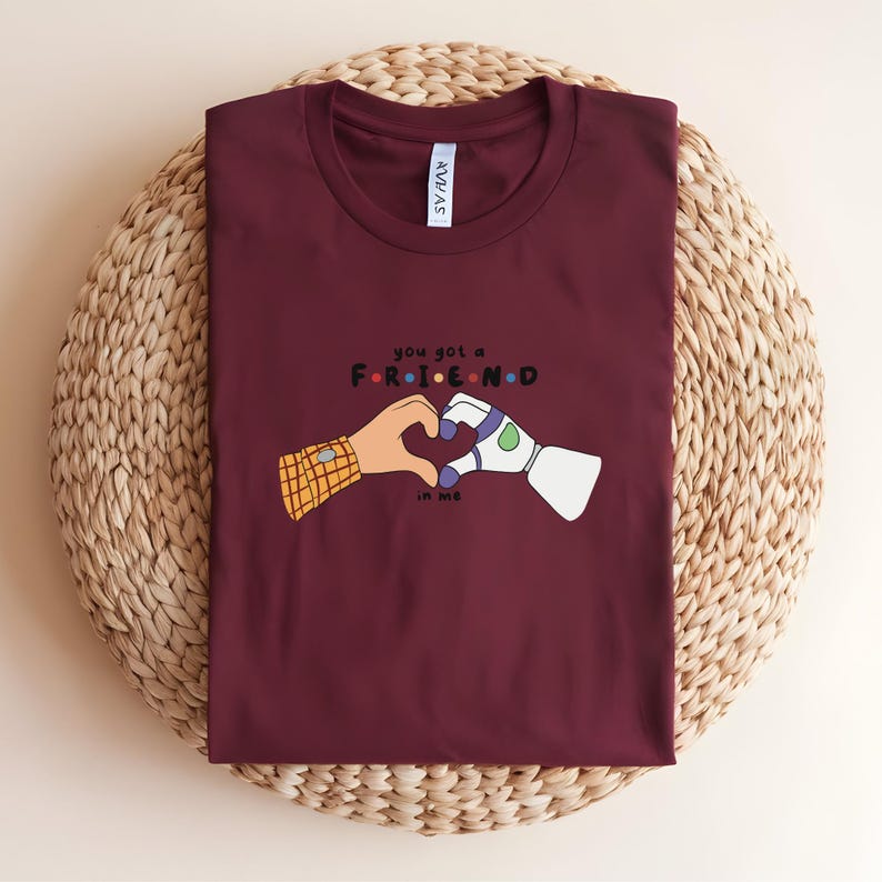 May include: A maroon t-shirt with the text "you got a FRIEND in me" in a playful font. The graphic features two cartoon hands forming a heart shape, one with a plaid sleeve and the other a robotic arm.