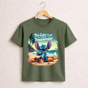 Pu&ograve; includere: T-shirt verde oliva con un personaggio dei cartoni animati su una tavola da surf sulla spiaggia. Il personaggio &egrave; blu con grandi orecchie e sorride. Il testo "Too Cute to be this Troublemaker" &egrave; sopra il personaggio.