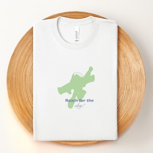 May include: White t-shirt with a green graphic of a figure in a flying pose and the text "Reach for the sky!" in blue. The shirt is folded and placed on a round, wooden tray.